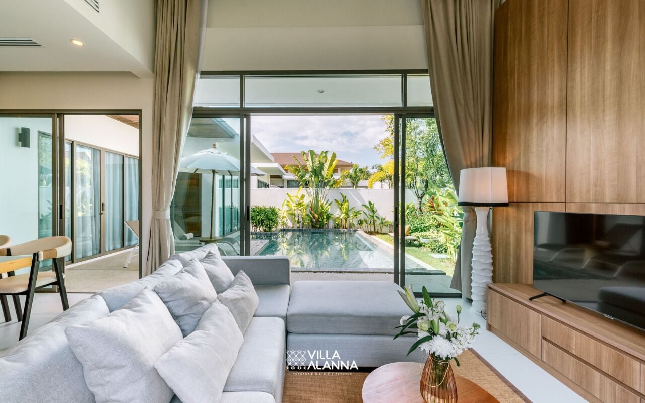 A modern living room with a grey sofa, a flat-screen TV, and a large window overlooking a pool and greenery.
