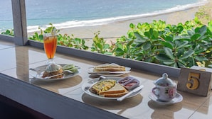 Ein Tisch mit Frühstücksangeboten und einem Teekessel, mit Blick auf einen Strand, das Meer und viel Grün.