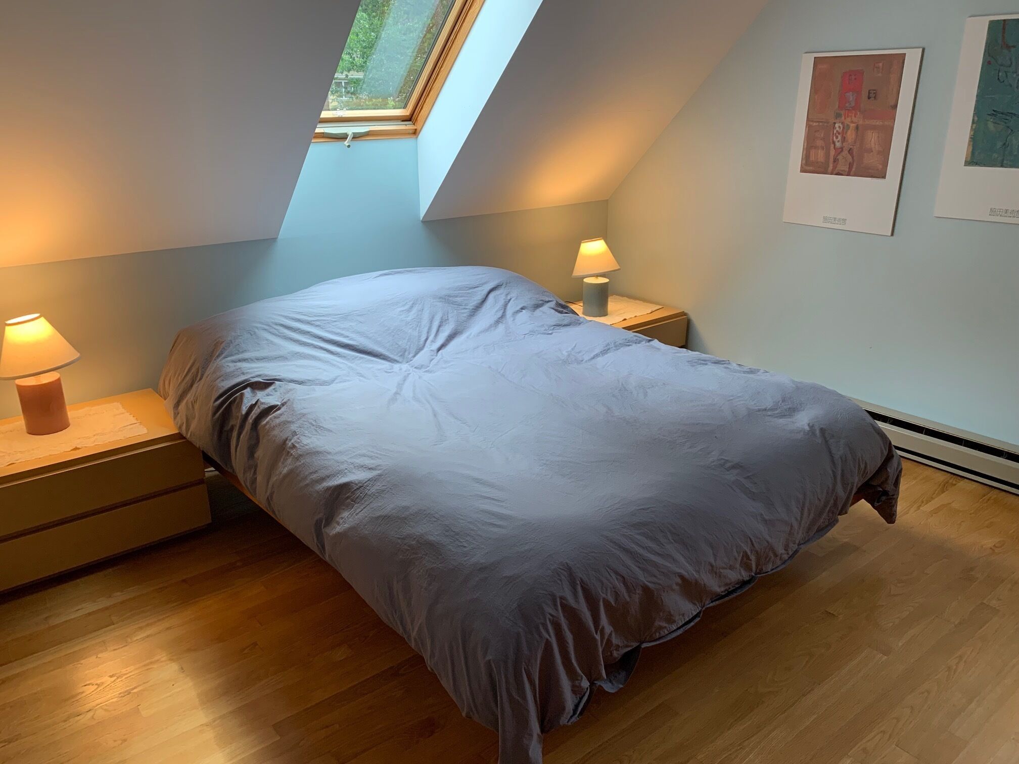 A bedroom with a skylight, a bed with a grey duvet, two bedside tables with lamps, and artwork on the wall.