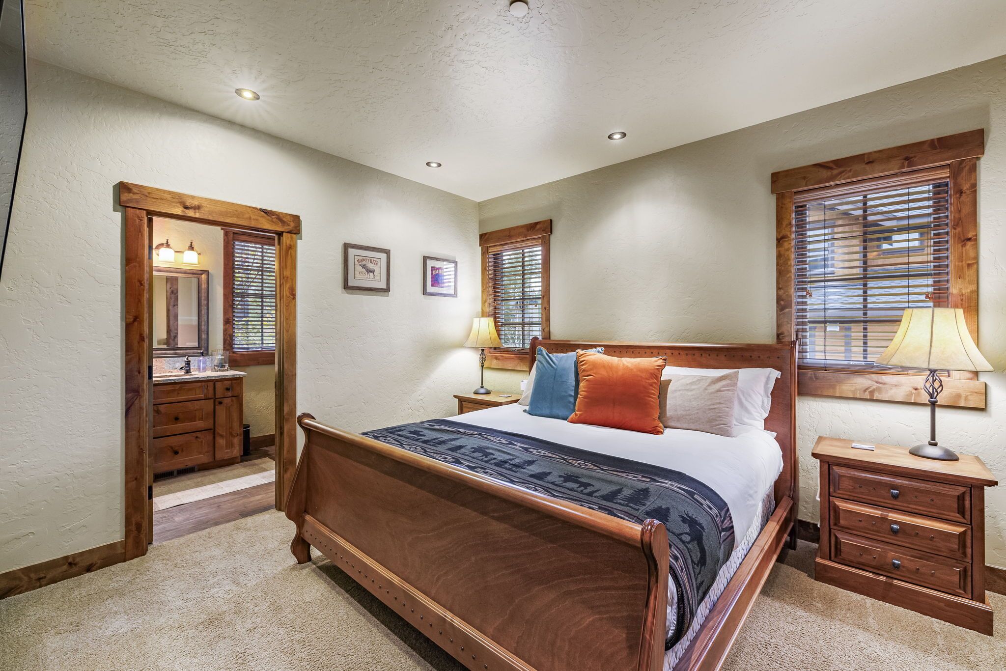 A bedroom with a wooden bed, two bedside tables, a nightstand, a mirror, and two windows with blinds.