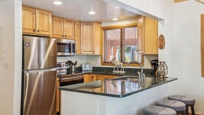 A modern kitchen with wooden cabinets, stainless steel appliances, a large island with stools, and a window with blinds.