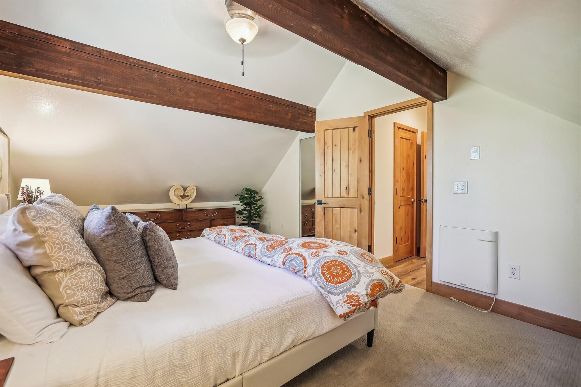 A bedroom with a bed, wooden beams, a ceiling light, a dresser, and a door leading to another room.