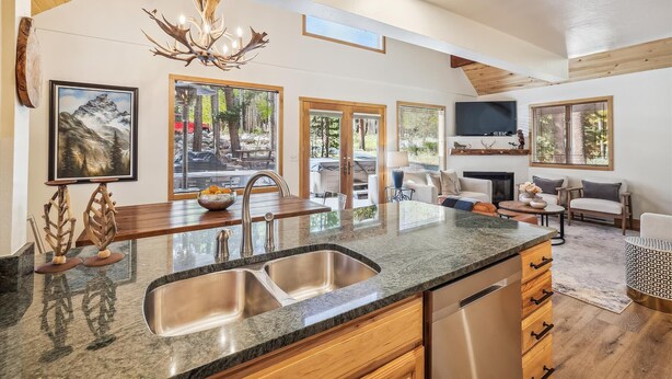 A modern kitchen with a granite countertop, stainless steel sink, and wooden cabinets. There is a dining area with a table and chairs, and a living room with a sofa and a fireplace.