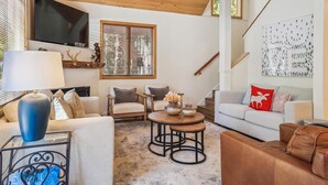 A cozy living room with a TV, comfortable seating, a fireplace, and a large window with blinds.