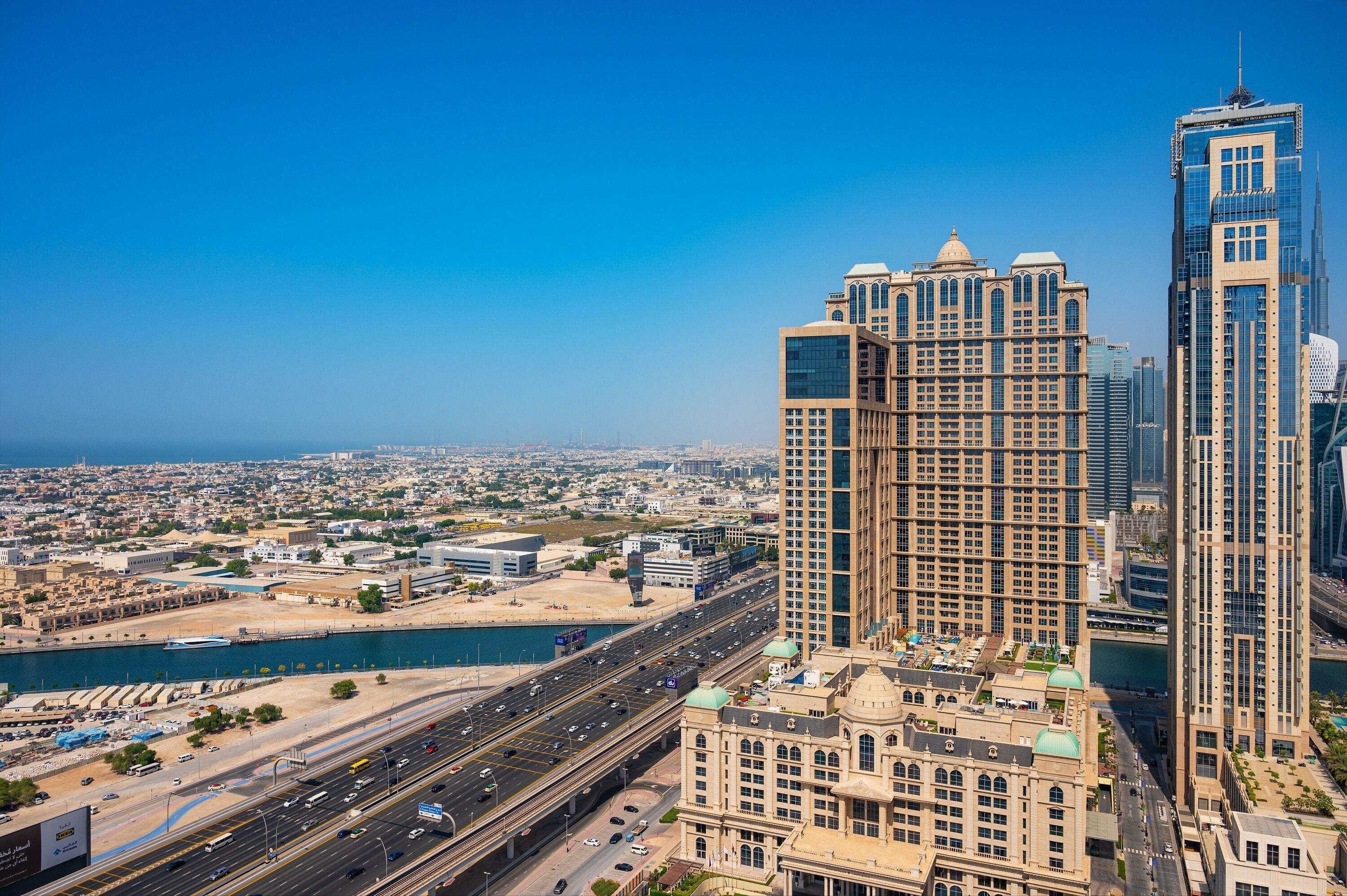 A high-rise building with a modern architectural design, situated near a body of water and a busy highway.