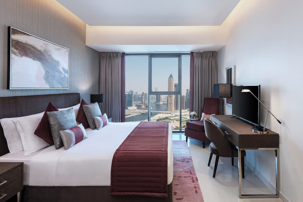 Ein modernes Hotelzimmer mit einem großen Bett, einem Schreibtisch, einem Fernseher und Blick auf eine Stadtsilhouette.