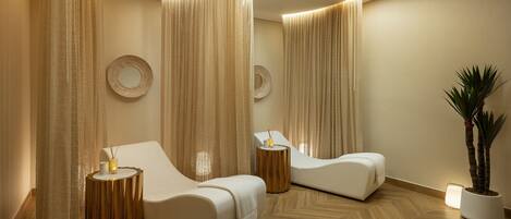 A modern spa room with two white lounge chairs, wooden side tables, and a potted plant.