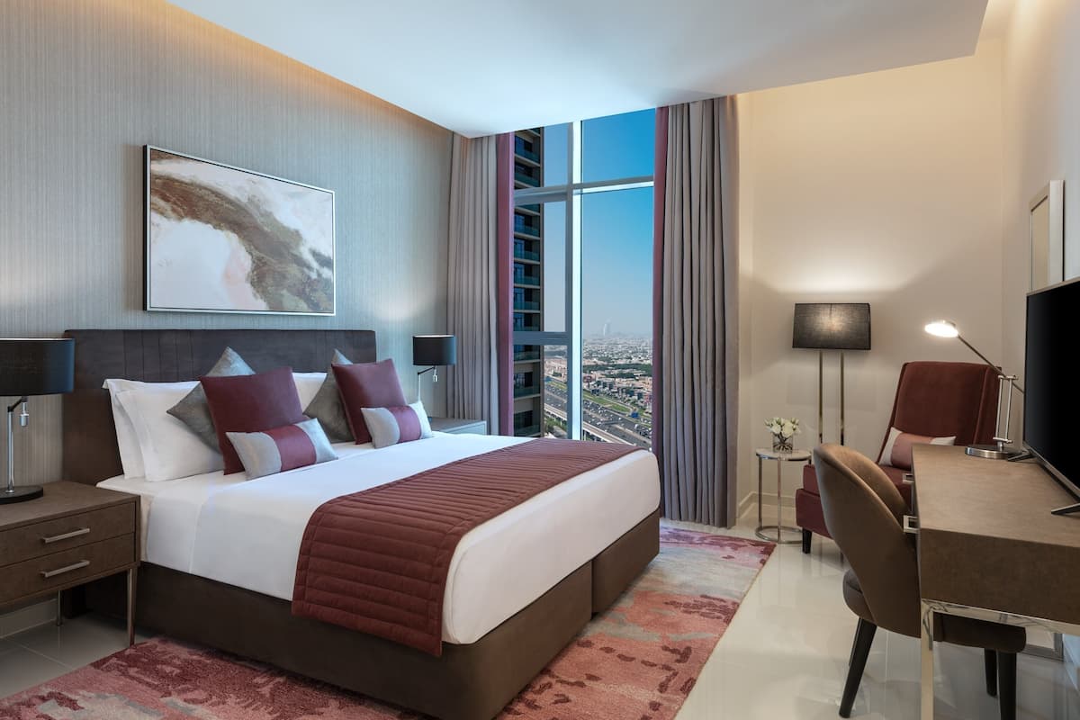 Ein modernes Hotelzimmer mit einem großen Bett, einem Schreibtisch, einem Sessel und Blick auf die Stadt durch große Fenster.
