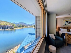 A room with a large window overlooking a lake and mountains, a bed with a wooden headboard, and a small table with a lamp.