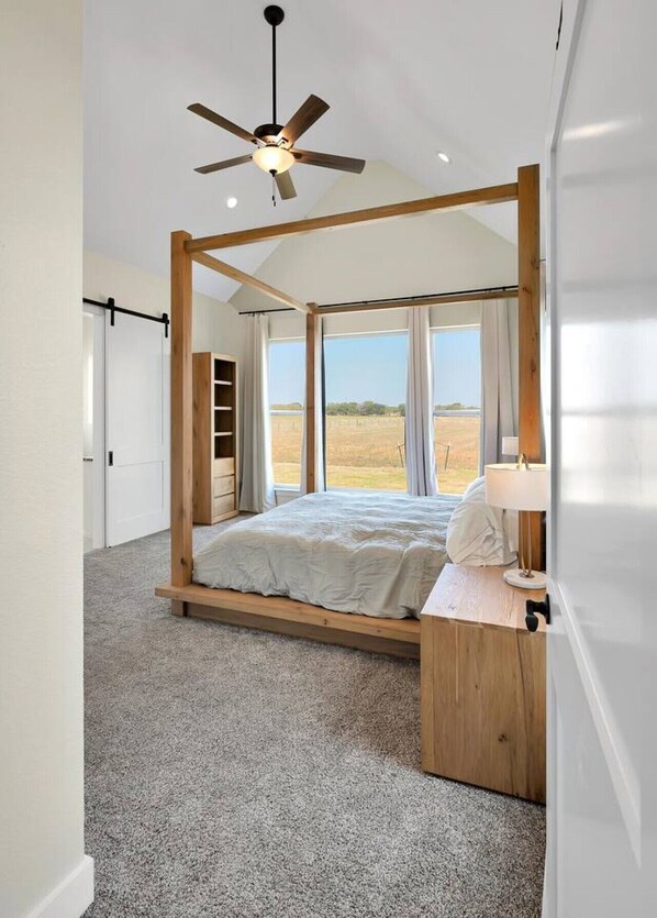 A bedroom with a four-poster bed, a ceiling fan, a desk with a lamp, and large windows offering a view of the outdoors.