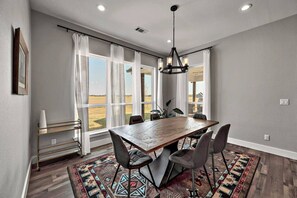 A dining area with a large wooden table, four chairs, a chandelier, and a view of the outdoors through large windows.