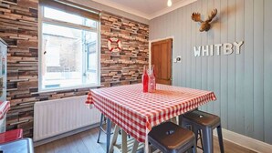 Un coin repas avec une table recouverte d’une nappe à carreaux rouges et blancs, deux tabourets, un mur en bois orné d’une décoration représentant un gilet de sauvetage et une tête de wapiti accrochée au mur.