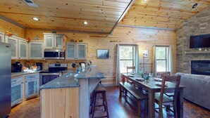 Interior rústico de una cabaña con paredes de madera, una cocina con encimeras de granito, una zona de comedor con una mesa y bancos de madera, y una chimenea de piedra.