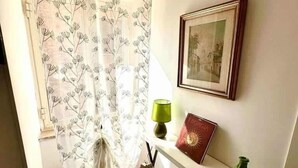 A room with a shower curtain, a framed picture on the wall, a small table with a lamp, and a chair.