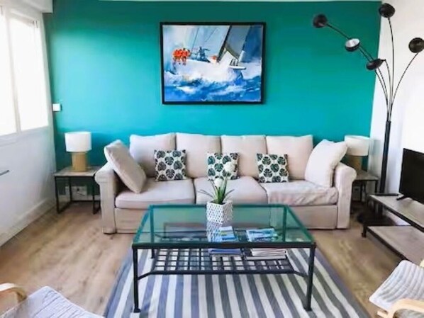 Un salon avec un mur de couleur turquoise, un canapé agrémenté de coussins à motifs, une table basse en verre et un tableau représentant un voilier.