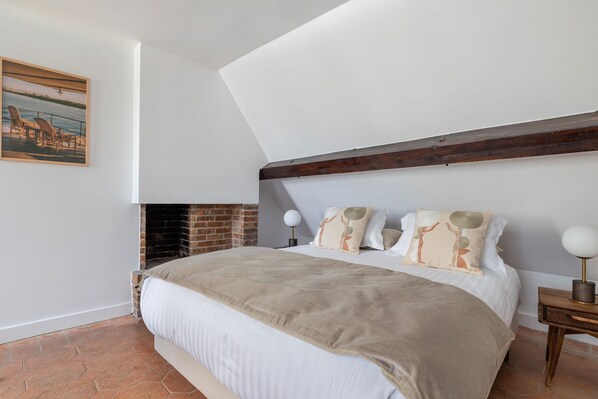 A bedroom with a sloped ceiling, a brick fireplace, a bed with a beige blanket, and a nightstand with a lamp. - Renaissance, 2BR/1BA, 5 people (París)