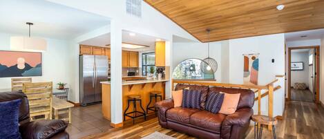A spacious living room with a brown leather sofa, a wooden dining table, and a kitchen area in the background.