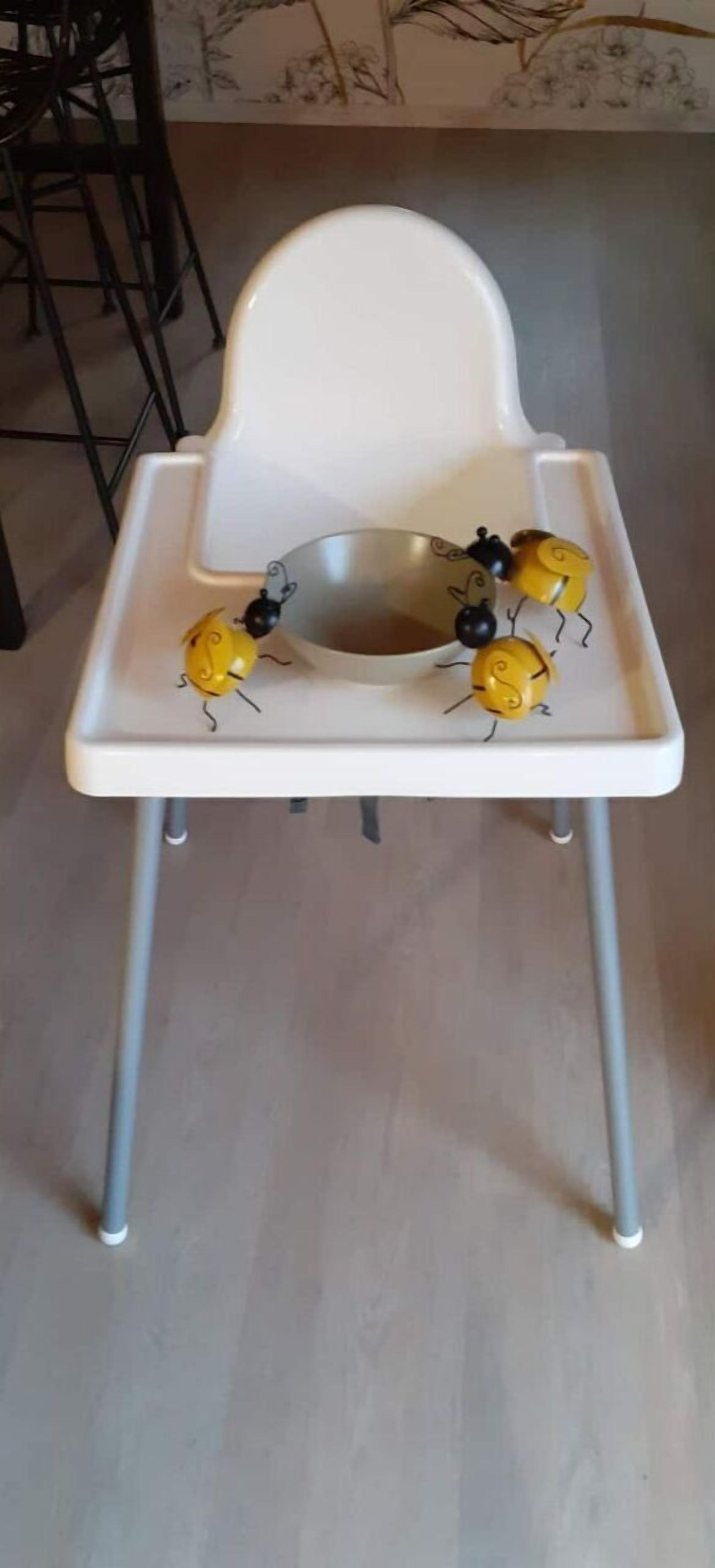 Ein weißer Hochstuhl mit einer Metallschalensitzfläche, vier gelb-schwarzen Spielzeugbienen und einem grauen Boden.