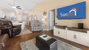 A living room with a TV displaying 'Sunset Nightly Rentals' logo, a leather sofa, a grey armchair, a dining area, and a kitchen in the background.