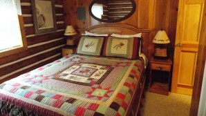 A wooden cabin bedroom with a large bed, two bedside tables with lamps, a round mirror above the bed, and a door to the right.