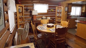 A cozy cabin interior with a wooden dining table, chairs, a fan, and a kitchen area with wooden cabinets and a window.