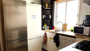 A compact kitchen with a stainless steel refrigerator, a microwave, and a dishwasher. There is a window above the sink, and various food items are stored on the shelves. - Anchor House Albufeira (Albufeira)