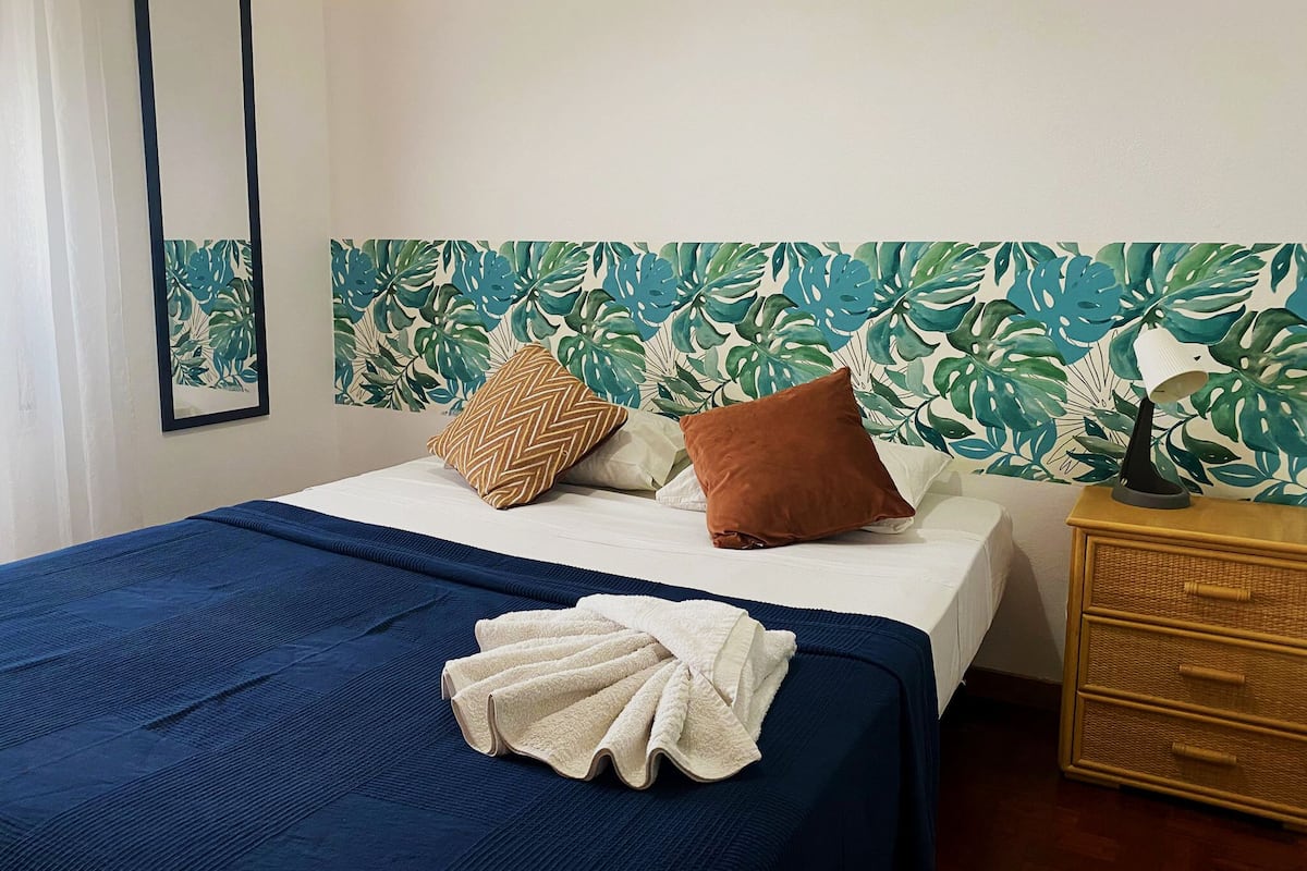A bed with a blue bedspread, two pillows, a white towel, and a nightstand with a lamp.