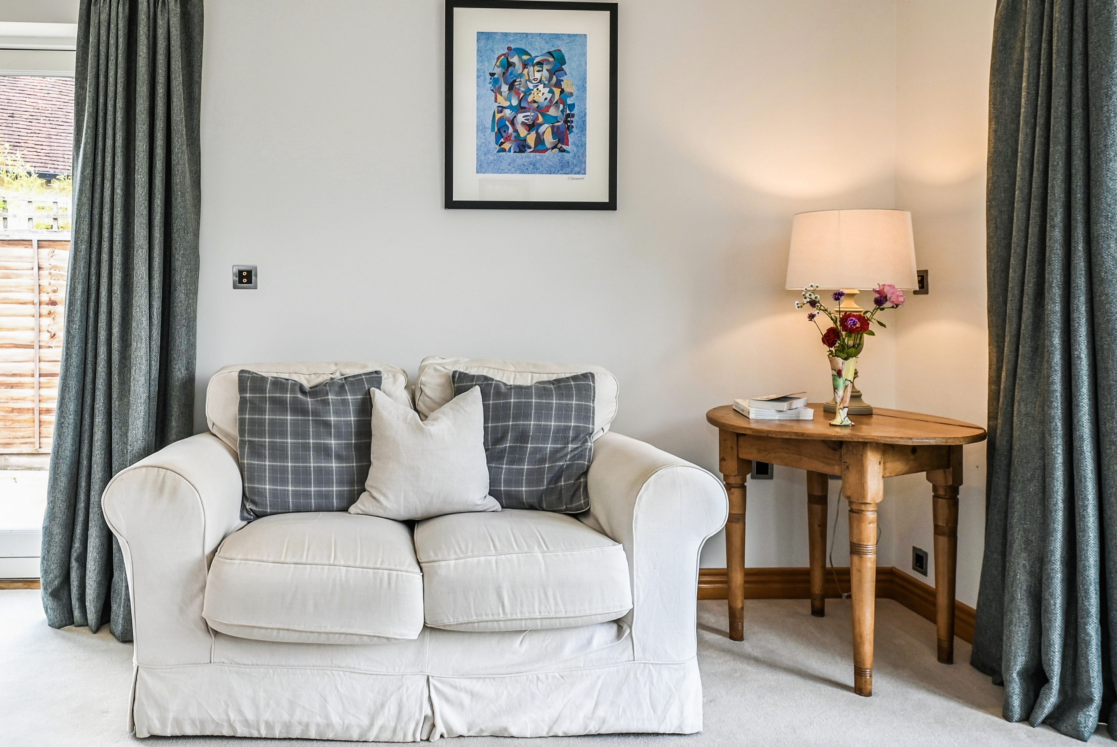 A white sofa with grey and white cushions, a framed abstract painting, a table with a lamp, and grey curtains.