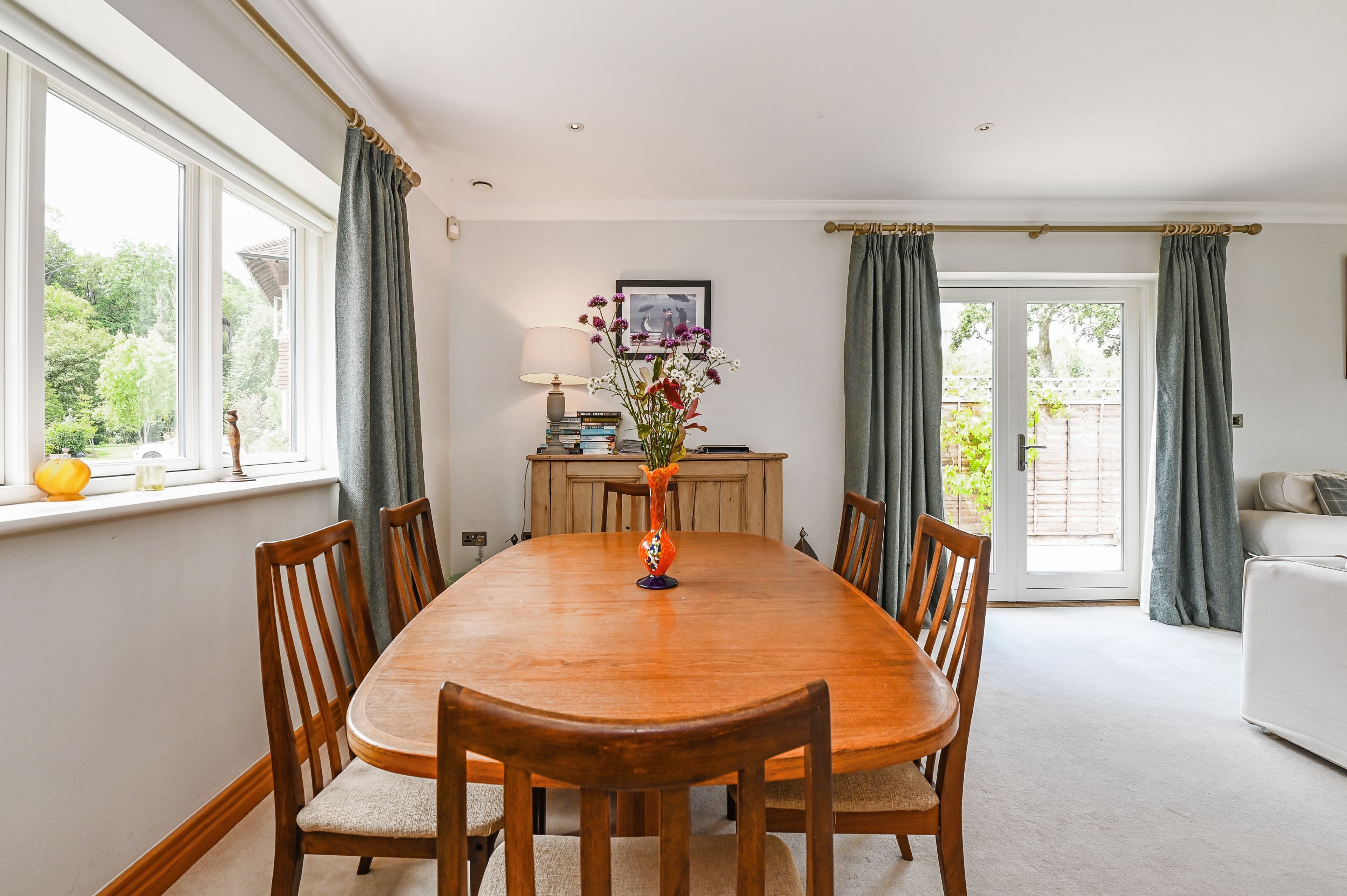 A dining area with a wooden table and chairs, a vase with flowers, a lamp, and a view of the outdoors through large windows.