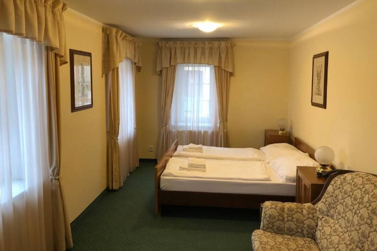A hotel room with a double bed, a chair, a bedside table with a lamp, and curtains on the windows.