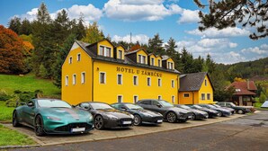 A hotel with a yellow facade, multiple cars parked in front, and a scenic backdrop of trees and hills. - Hotel U Zámečku Cihelny (Karlovy Vary)