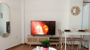 Una sala de estar moderna con un televisor de pantalla plana, un comedor con mesa y sillas, y una pequeña mesa redonda blanca con una planta en maceta y un objeto decorativo.