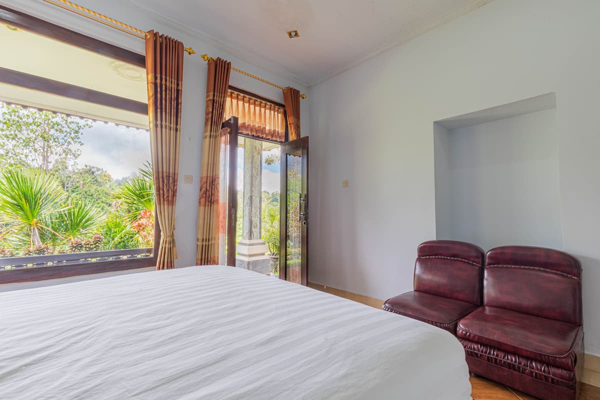 A bedroom with a large bed, a pair of leather chairs, a sliding glass door leading to a balcony with a view of greenery.