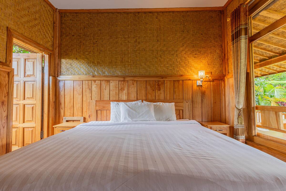 A wooden-paneled room with a large bed, a nightstand, and a window with a view of greenery.