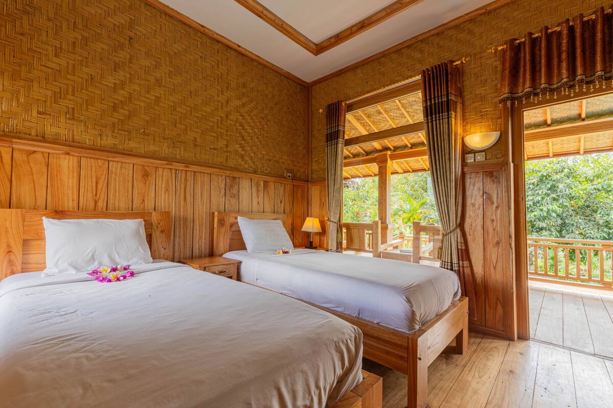 A room with two wooden beds, a wooden ceiling, and a large window with curtains.