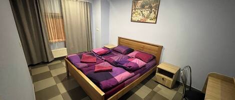 A bedroom with a checkered floor, a wooden bed with purple bedding, a nightstand, a chair, and a painting on the wall.