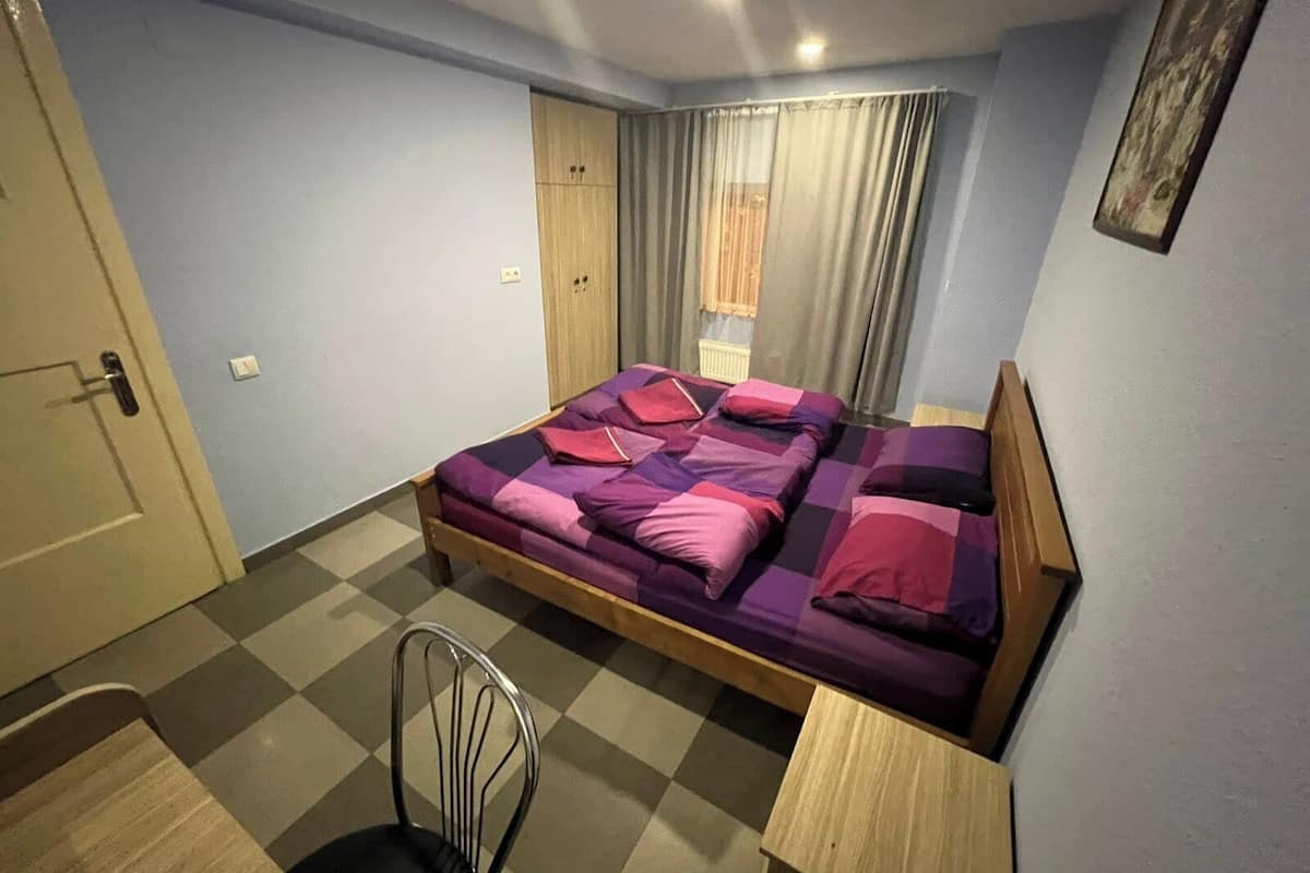 A bedroom with a bed, a chair, a wooden nightstand, a wardrobe, and a window with curtains.