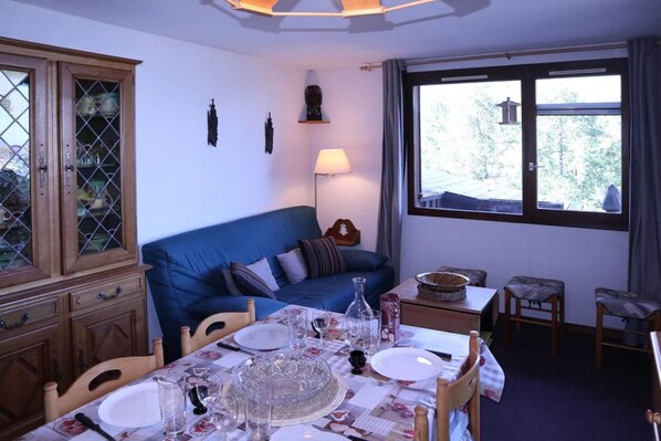 A dining area with a table set for two, a blue sofa, a cabinet with glass doors, and a window with a view of trees.