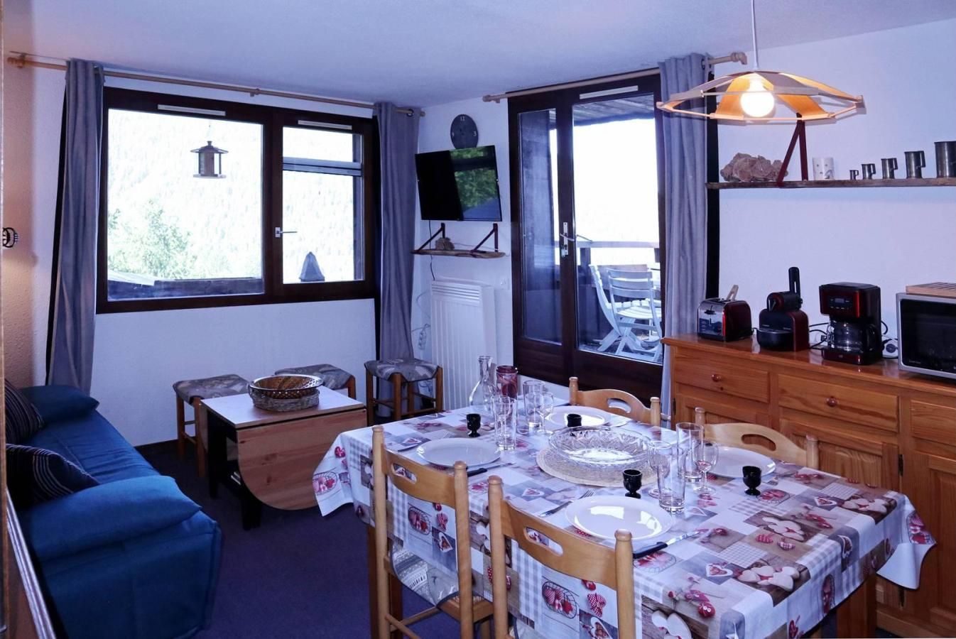 A dining area with a table set for four, a television on the wall, and a kitchen area with appliances.