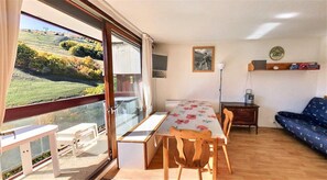 Un espace repas comprenant une table et des chaises, un canapé et une vue sur un paysage verdoyant, visible à travers une grande fenêtre.