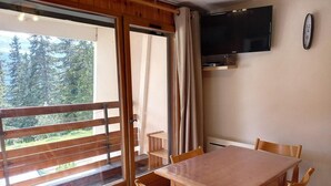 Een kamer met een houten tafel en stoelen, een aan de muur bevestigde televisie en een schuifdeur naar een balkon met uitzicht op bomen.
