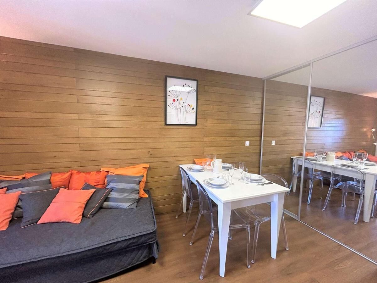 A room with a wooden wall, a bed with orange and gray pillows, a white table with chairs, and a large mirror reflecting the room.