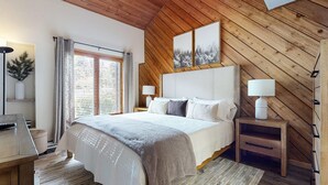 A bedroom with a wooden ceiling, a large bed with white bedding, two bedside lamps, a window with curtains, and a framed picture above the bed.