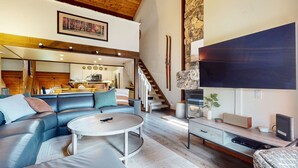 A spacious living area with a leather sofa, a round coffee table, and a flat-screen TV. A kitchen is visible in the background, and there is a staircase leading to another level.