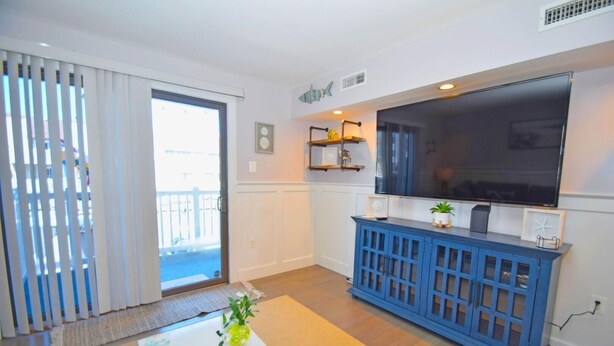 Una sala de estar con un televisor grande, un mueble azul y un ventanal corredizo que da a un balcón.