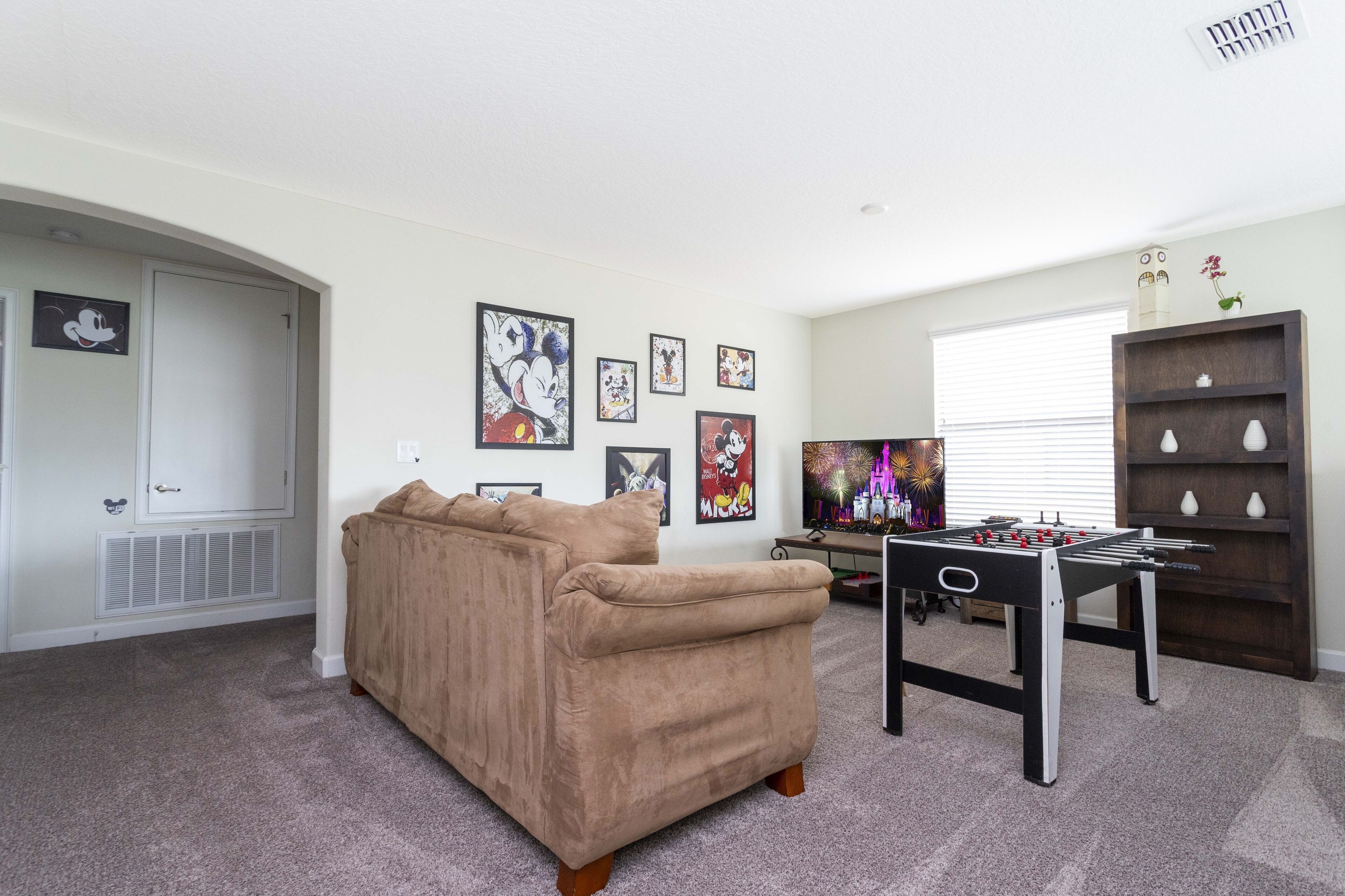 A living room with a beige sofa, a foosball table, and wall art featuring cartoon characters.