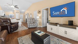 Een woonkamer met een televisie waarop 'Sunset Nightly Rentals' staat, een bruine leren bank, een grijze fauteuil, een klein wit tafeltje en een kast.