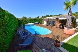 Una terraza de madera con piscina, mobiliario exterior y una pequeña construcción con techo de paja.