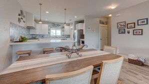 Une cuisine moderne avec des meubles blancs, un îlot central, des luminaires suspendus et un coin repas comprenant une table et des chaises en bois.