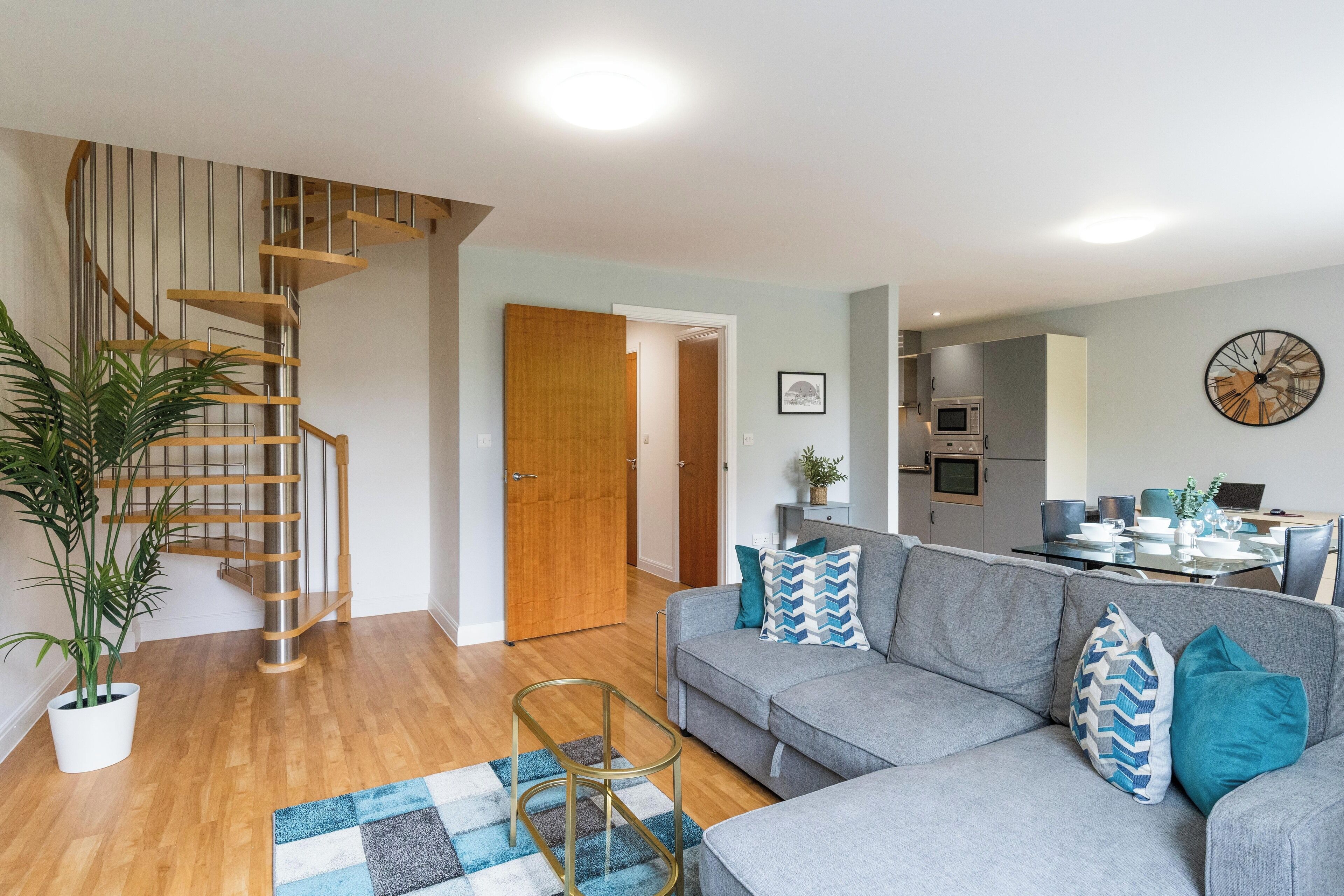 Ein modernes Wohnzimmer mit grauem Sofa, einer Holztreppe und einem Essbereich mit Tisch und Stühlen.